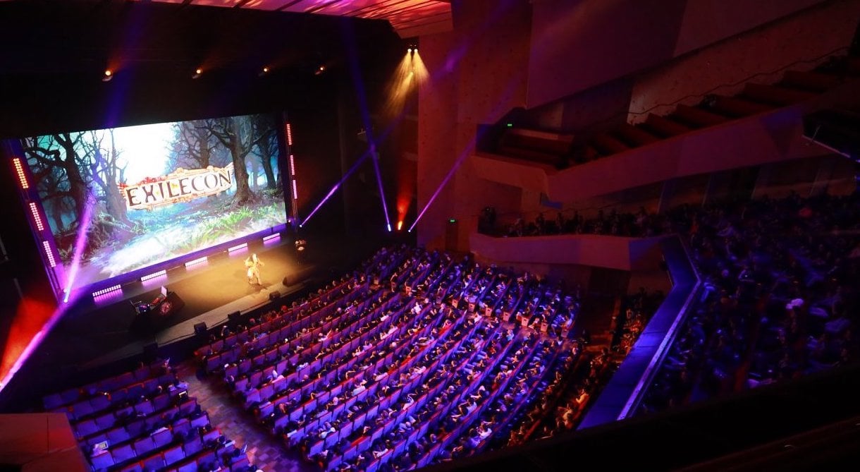 A large, dark conference room with the words "ExileCon" written on the  projector screen.